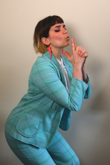 Comedian Kate Hammer strikes a playful pose in a blue plaid suit, representing the vibrant Monkey Barrel comedy scene.