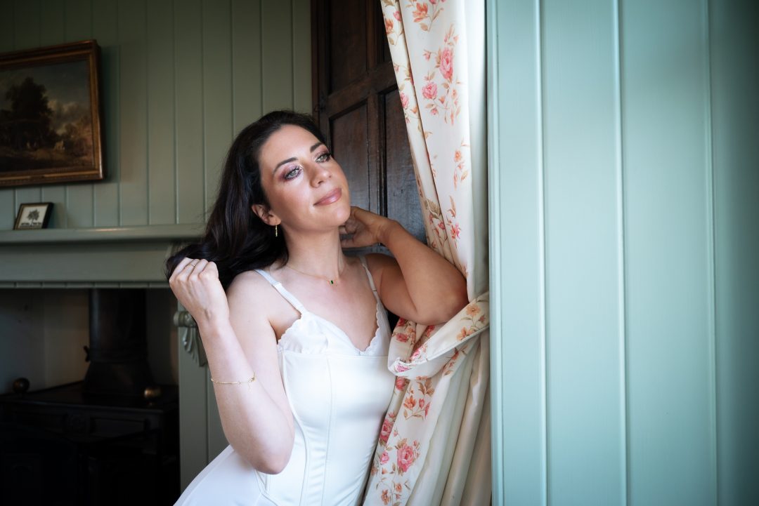 Musician Kohla in a white corset top posing beside a floral curtain