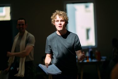 Actor Jude Bain holding script pages during a rehearsal with another cast member blurred in the background.