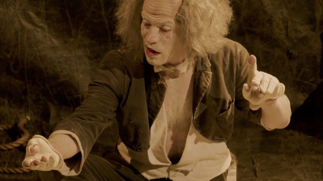 A performer in a theatrical costume with white face makeup, a bald cap, and wild grey hair, gesturing with white-gloved hands against a dark background.