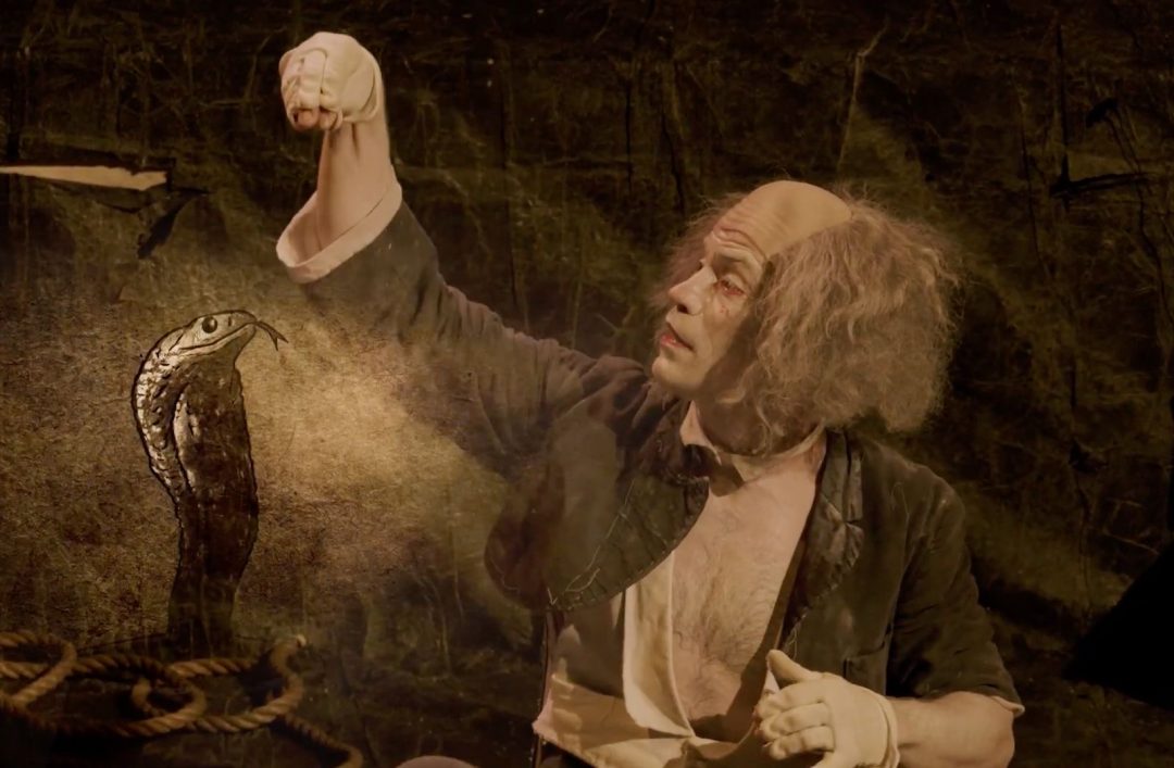 A theatrical performer with white face makeup and wild grey hair, holding up a gloved hand next to an animated illustration of a cobra snake.
