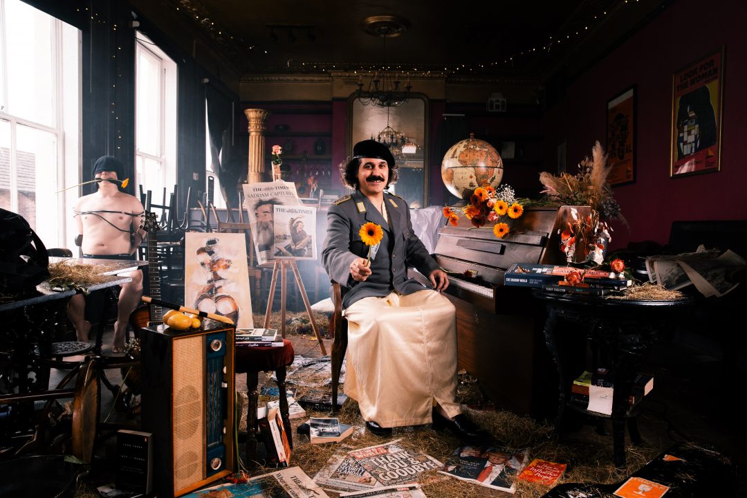 Sami Abu Wardeh sitting in a dimly lit, chaotic room holding out a yellow flower. He wears a grey military-style jacket.