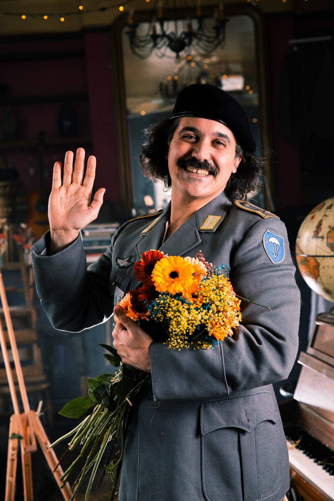 Sami Abu Wardeh smiling and waving at the camera while holding a bright bouquet of flowers. He is wearing a grey military-style jacket with a blue patch and a black beret.