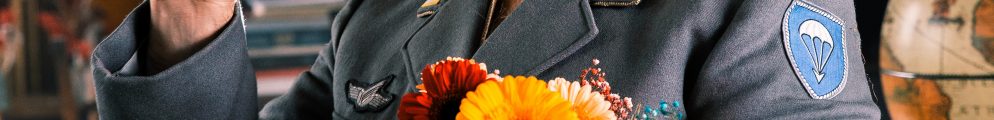 Sami Abu Wardeh smiling and waving at the camera while holding a bright bouquet of flowers. He is wearing a grey military-style jacket with a blue patch and a black beret.