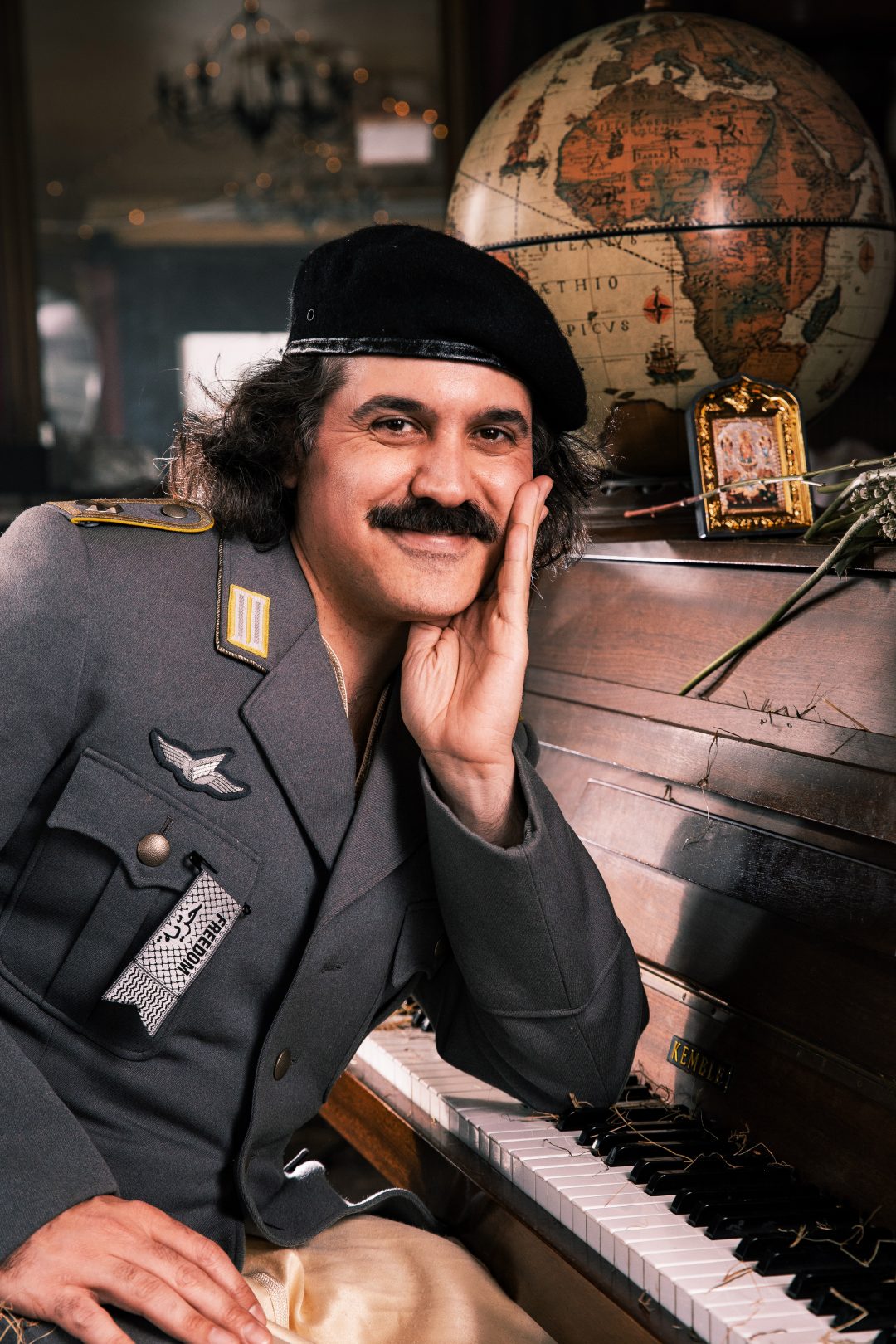 A smiling portrait of Sami Abu Wardeh leaning his cheek against his hand on an old wooden piano. He is wearing a black beret and a grey military-style jacket featuring a patch that reads 'Freedom'.