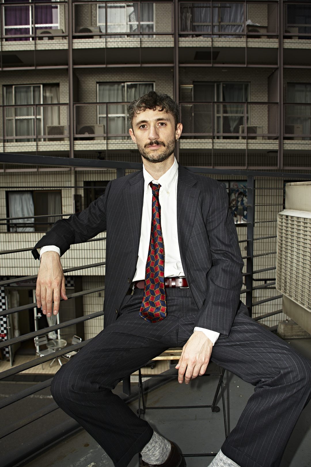 Steven Phillips-Horst sitting on a metal stool outdoors in a dark pinstripe suit, white button-down shirt, and patterned red and blue tie against a balcony and building backdrop for The Last Mad Man.