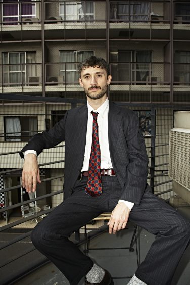Steven Phillips-Horst sitting on a metal stool outdoors in a dark pinstripe suit, white button-down shirt, and patterned red and blue tie against a balcony and building backdrop for The Last Mad Man.