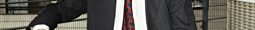 Steven Phillips-Horst sitting on a metal stool outdoors in a dark pinstripe suit, white button-down shirt, and patterned red and blue tie against a balcony and building backdrop for The Last Mad Man.