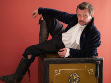 Comedian Thom Tuck in character as Sir Gregor MacGregor for the Edinburgh Fringe show The Wolf of Poyais, lounging on a vintage John Tann's Reliance safe against a red background.