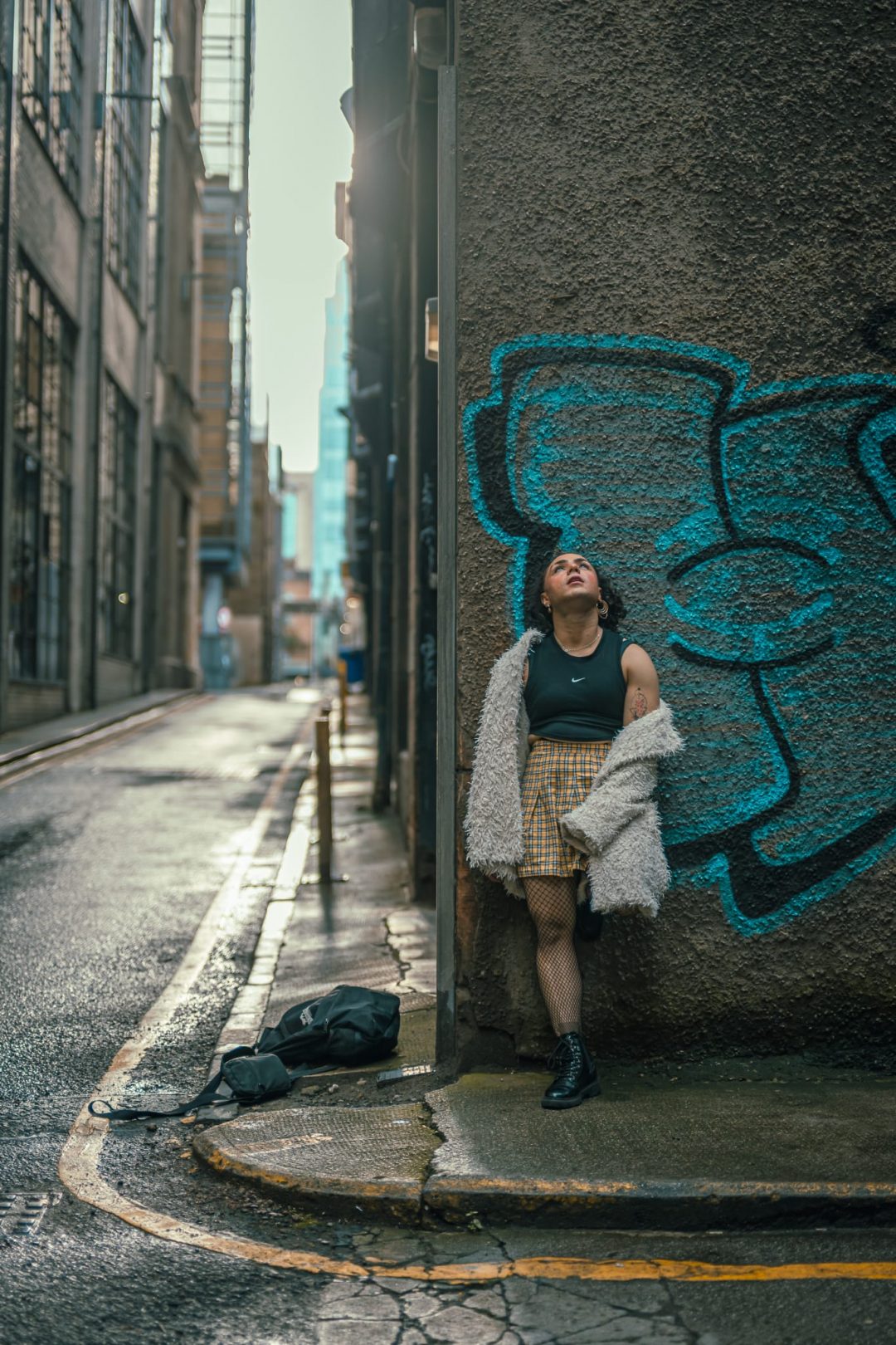 Comedian Tia Rey leaning against a textured brick wall with blue graffiti in an alleyway, wearing a black crop top, yellow plaid skirt, fishnets, and a fluffy white jacket, looking upward.
