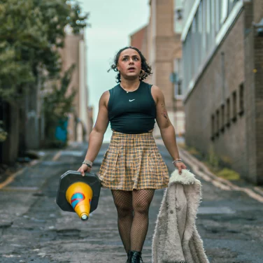 Comedian Tia Rey walking down an urban alleyway carrying a traffic cone in one hand and a fluffy white jacket in the other, wearing a black crop top, yellow plaid skirt, fishnets, and boots.
