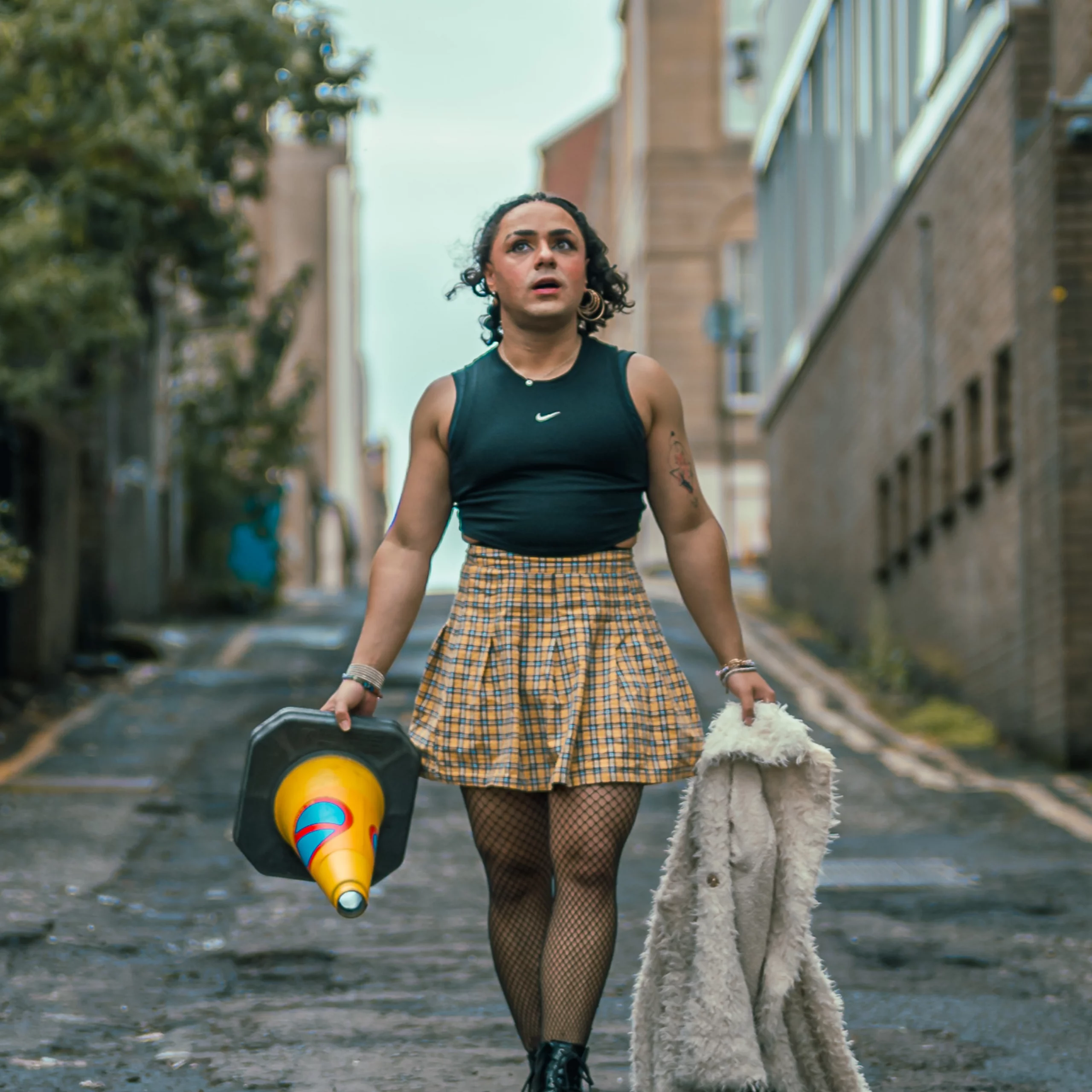 Comedian Tia Rey walking down an urban alleyway carrying a traffic cone in one hand and a fluffy white jacket in the other, wearing a black crop top, yellow plaid skirt, fishnets, and boots.