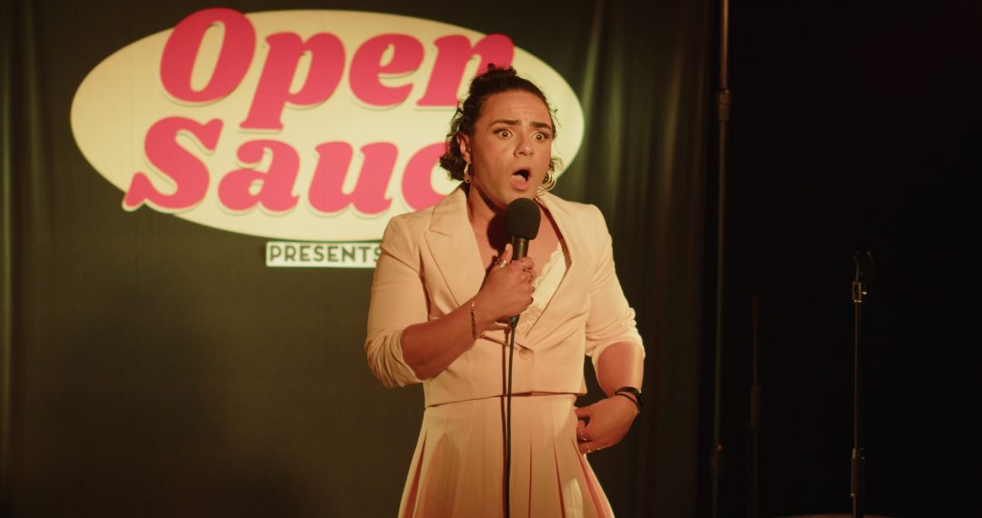 Comedian Tia Rey performing live on stage with a microphone in front of an 'Open Sauce Presents' backdrop, wearing a pink blazer and skirt.