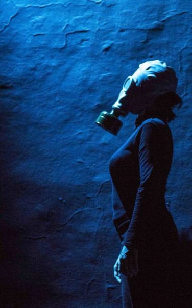 Elena Arvigo wearing a gas mask in profile against a textured blue wall in the stage production Atomic Tales.