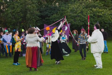 Costumed performers dance with ribbons on a grassy lawn during the Sett Studios Foik Ferly procession.