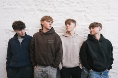 Four members of indie band Blanc standing side by side against a white textured stone wall.