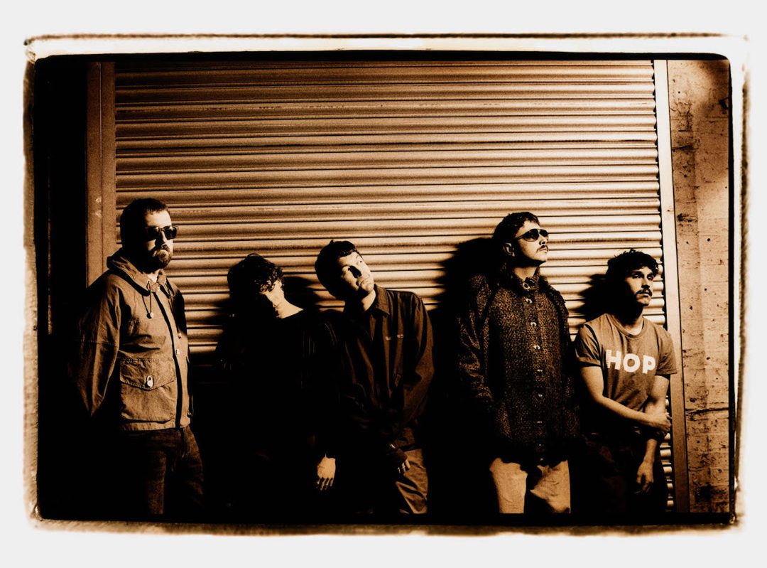 Sepia-toned, vintage Polaroid-style photograph of Gurriers leaning against a distressed urban roller shutter, featuring a white border.